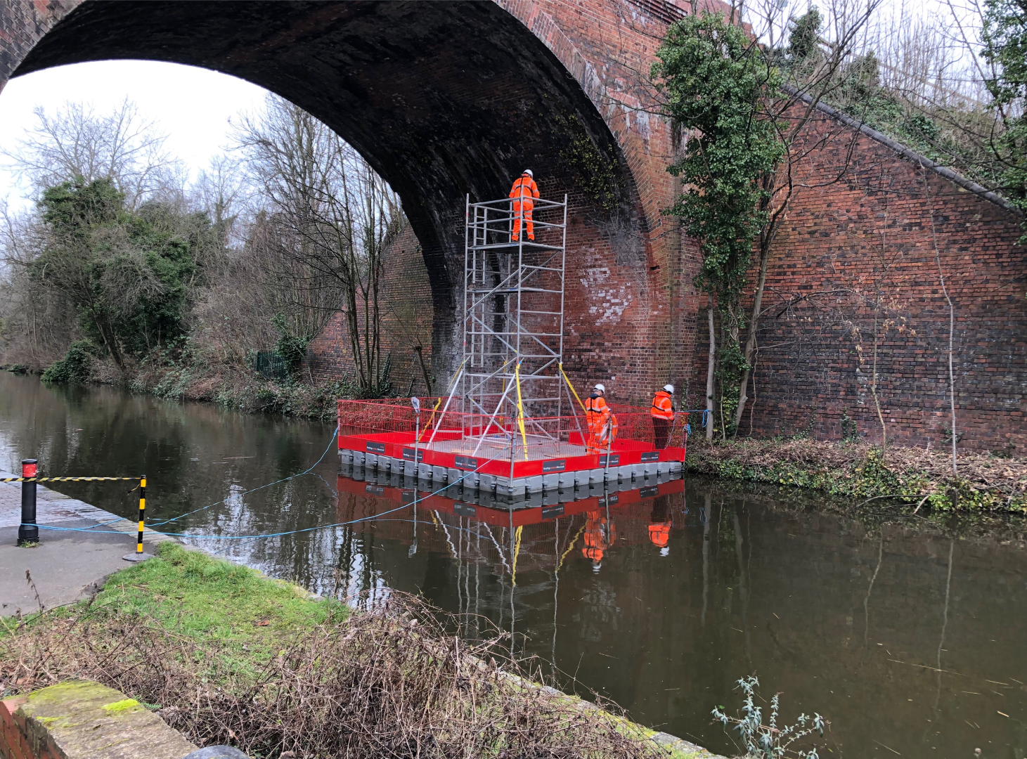 Floating pontoon work platform supporting scaffold access for inspection and maintenance beneath a brick arch bridge
