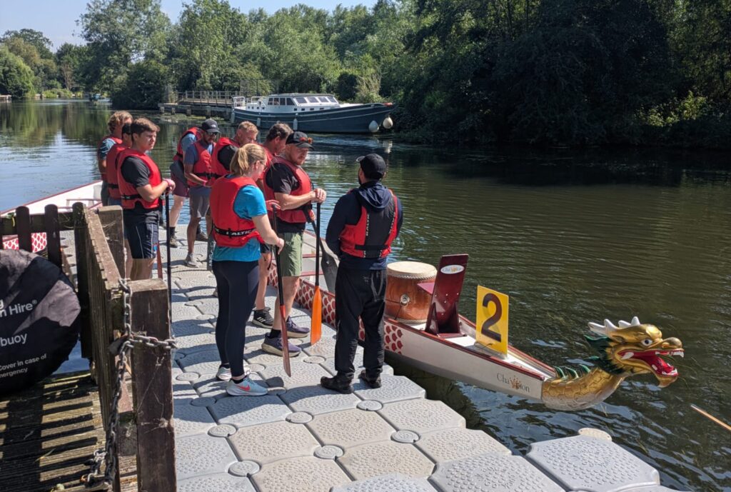 A team in red life jackets stands on a modular floating pontoon preparing to board a dragon boat with paddles, on a tree-lined river.