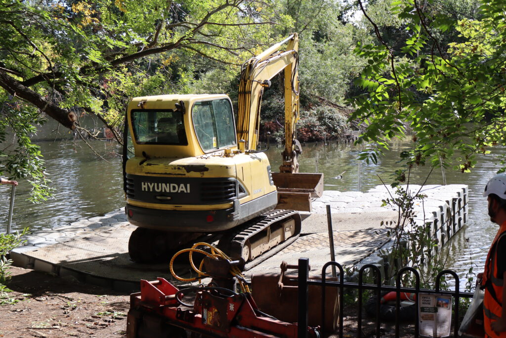 Tracked excavator operating on a double-stacked modular floating pontoon beside a wooded riverbank.